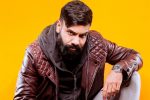 Paul Chowdhry sat down with his left arm on one knee wearing a brown hooded jacket, red leather jacket, grey jeans and a flashy watch with a yellow backdrop